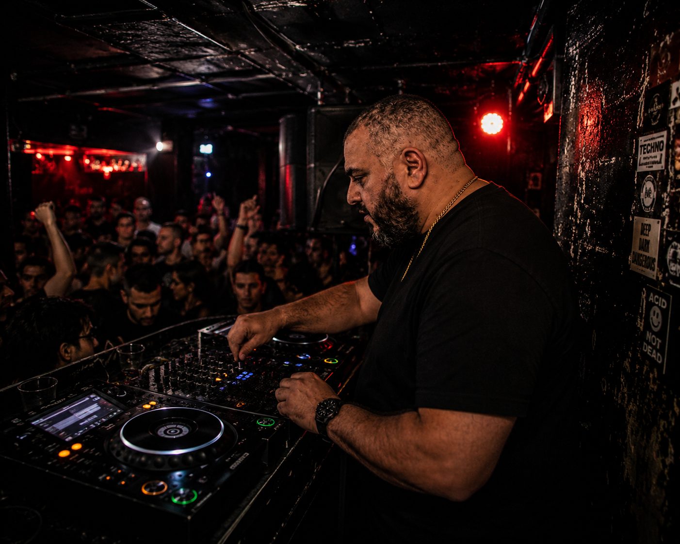 Adam J. Colosimo DJing in a dark underground club lit in red, crowd visible behind